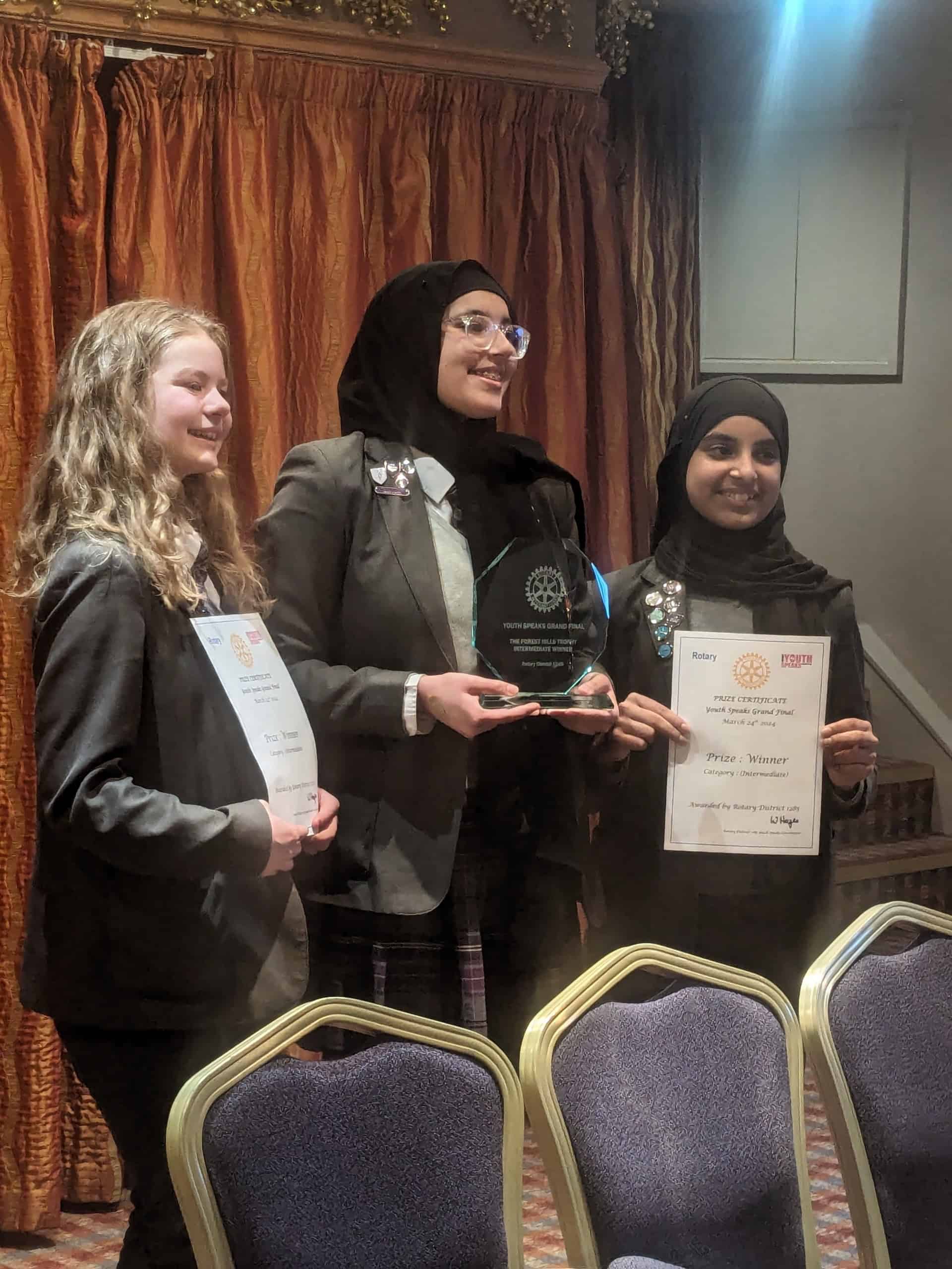 Three students from Laurus Cheadle Hulme smile as they hold their certificates and trophy which they received as the Intermediate Winners of the Rotary Youth Speaks District 1285 Grand Final 2024.